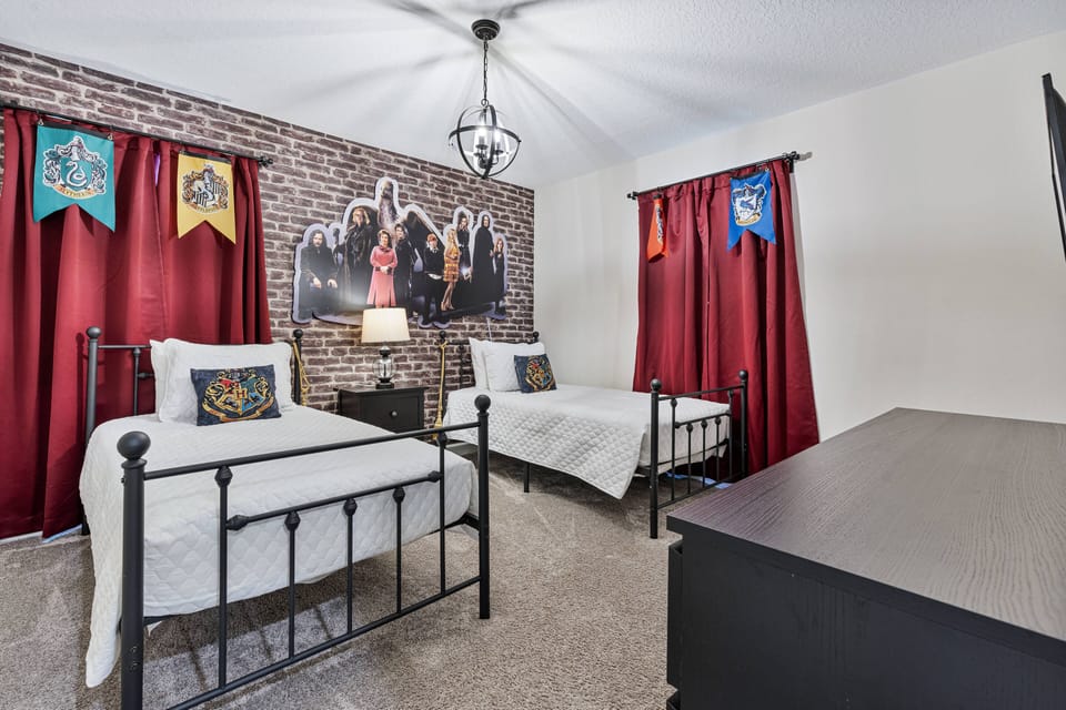 Experience a charming bedroom with two stylish metal beds, bright accent pillows, rich red curtains, and a striking brick wall that adds character, all illuminated by a warm lamp for a cozy atmosphere.