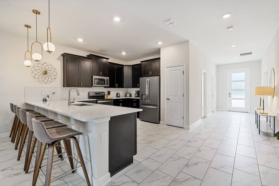 Experience a stylish kitchen with modern dark cabinetry and a spacious island with high-backed stools, ideal for casual dining. The polished tile floor shines under chic pendant lights, creating a warm and inviting ambiance.
