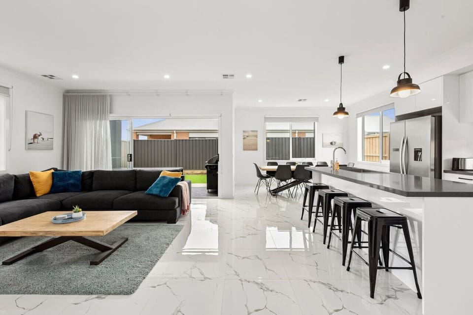 This spacious open-plan living area features a large sectional lounge, a modern dining setting, and a sleek kitchen with island seating. This space is perfect for entertaining or relaxing with family and friends