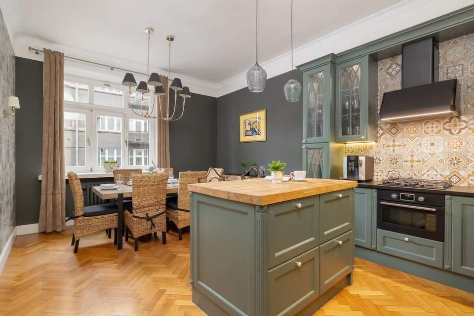 A charming kitchen with green cabinets, wooden countertops, and elegant glass doors. There’s a small dining table with chairs.