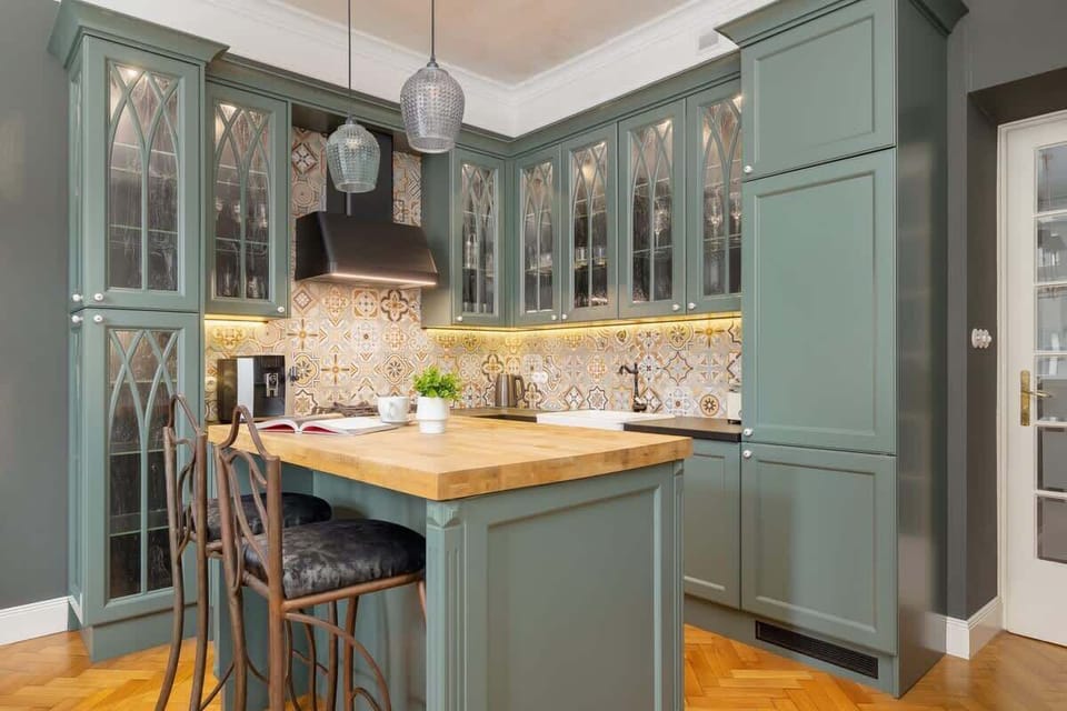 A charming kitchen with green cabinets, wooden countertops, and elegant glass doors. There’s a small dining table with chairs.