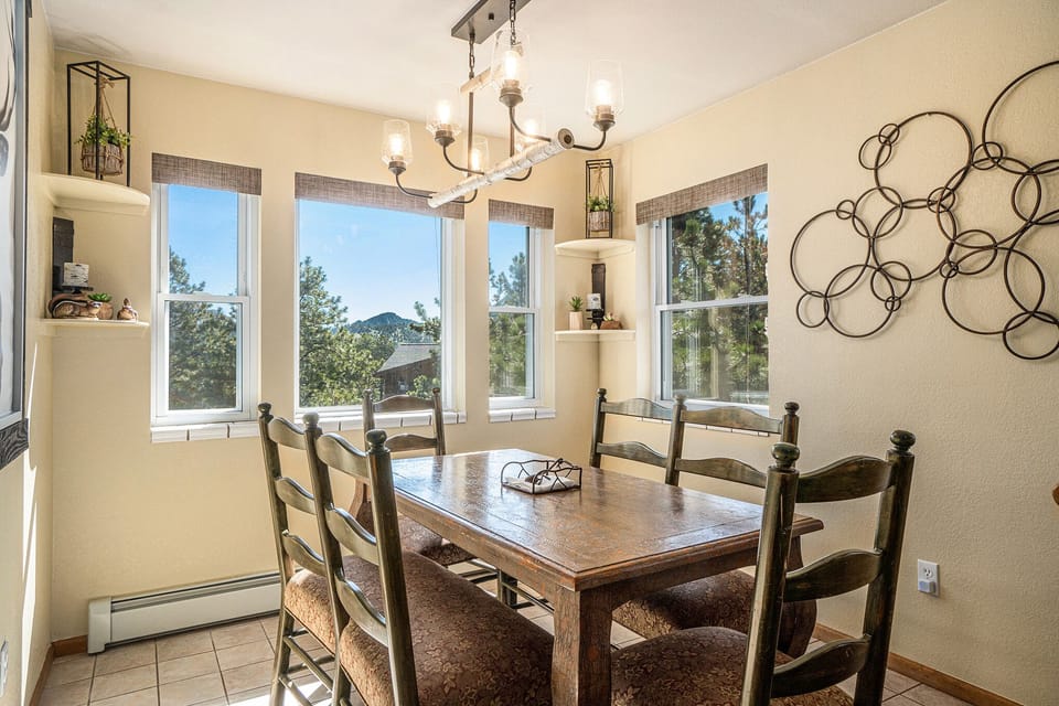 A dining room with a wooden table and six chairs. Large windows with a view of trees, shelves with decor, and a circular wall art piece are visible. A chandelier hangs above the table.