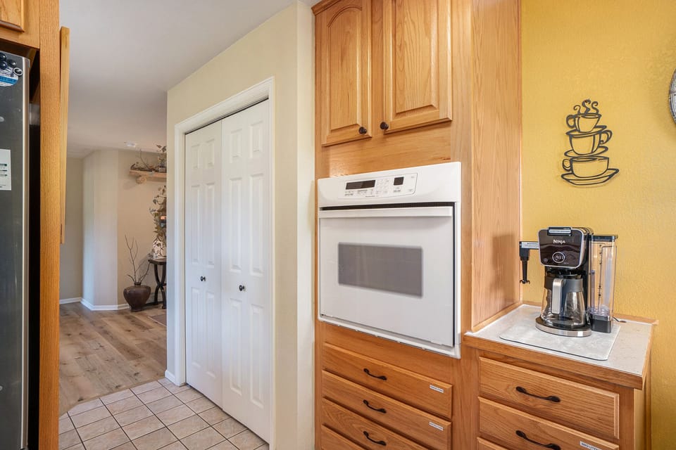 Kitchen corner with a built-in oven, wooden cabinets, and a countertop holding a coffee maker. White closet doors are in view, and a hallway leads to another room.