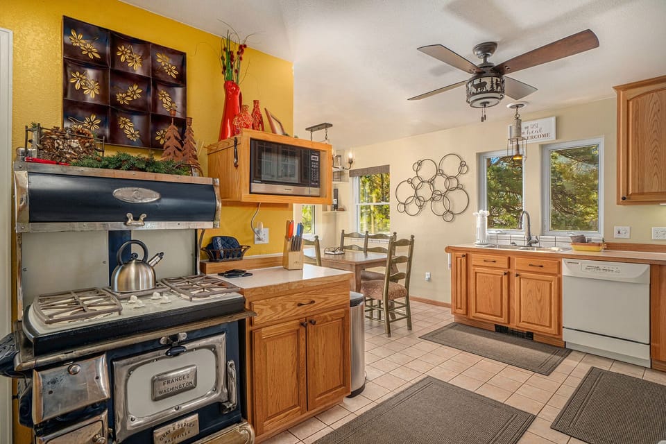 A cozy kitchen featuring a vintage stove, wooden cabinetry, colorful wall decor, ceiling fan, and dining area with windows in the background.