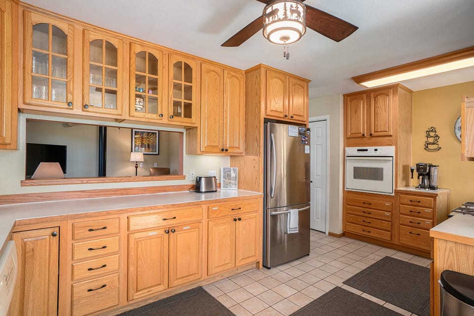 A spacious kitchen with wooden cabinets, a stainless steel refrigerator, built-in oven, and tile flooring. Ceiling fan with light fixture above. Counter appliances and decor are visible.