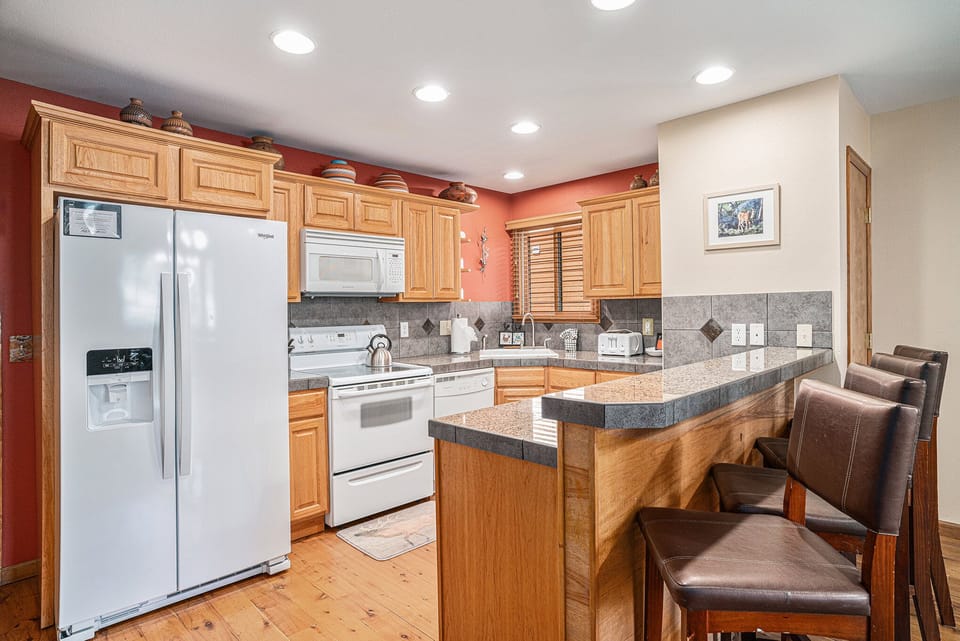 A kitchen with wooden cabinets, a white fridge, stove, microwave, and brown bar stools by a counter. There's a window with blinds and decorative pots on top of the cabinets.
