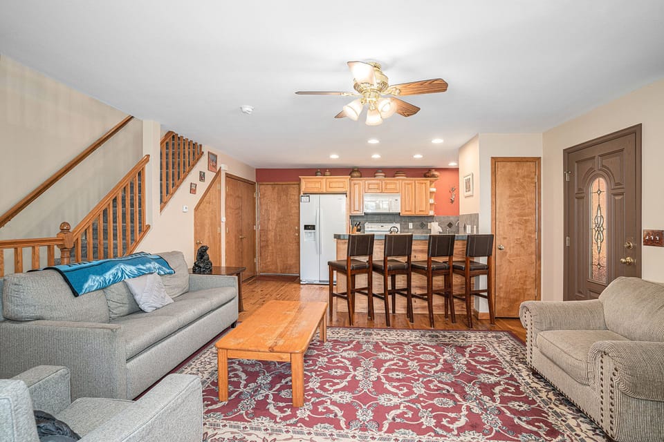Living room with gray sofas, wooden coffee table, patterned rug, ceiling fan, bar stools, and view of a kitchen with wooden cabinets and a refrigerator. Stairs lead to an upper floor.