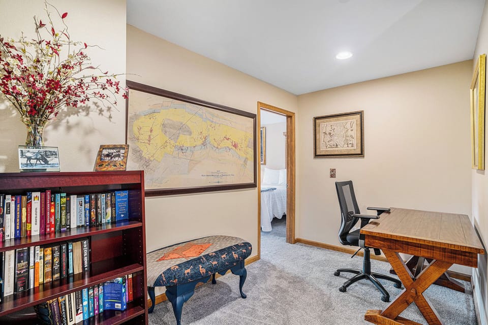 A home office with a wooden desk, black chair, and bookshelf. Wall maps and framed art decorate the beige walls. A floral arrangement sits atop the bookshelf. Carpeted flooring is visible.