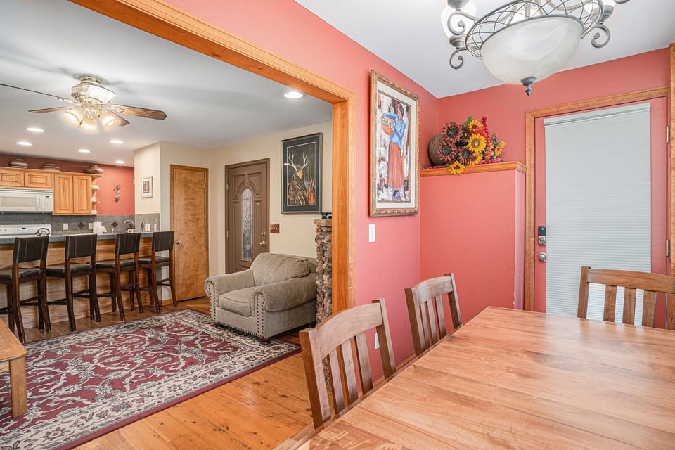 A cozy dining area with a wooden table and chairs. Adjacent, an open kitchen features bar stools, a ceiling fan, and artwork on red walls. A door with blinds is in the corner.
