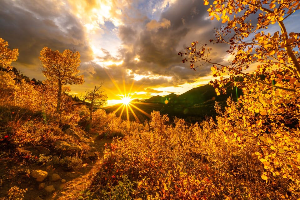 Golden autumn trees in a mountainous landscape at sunset, with sun rays streaming through clouds.