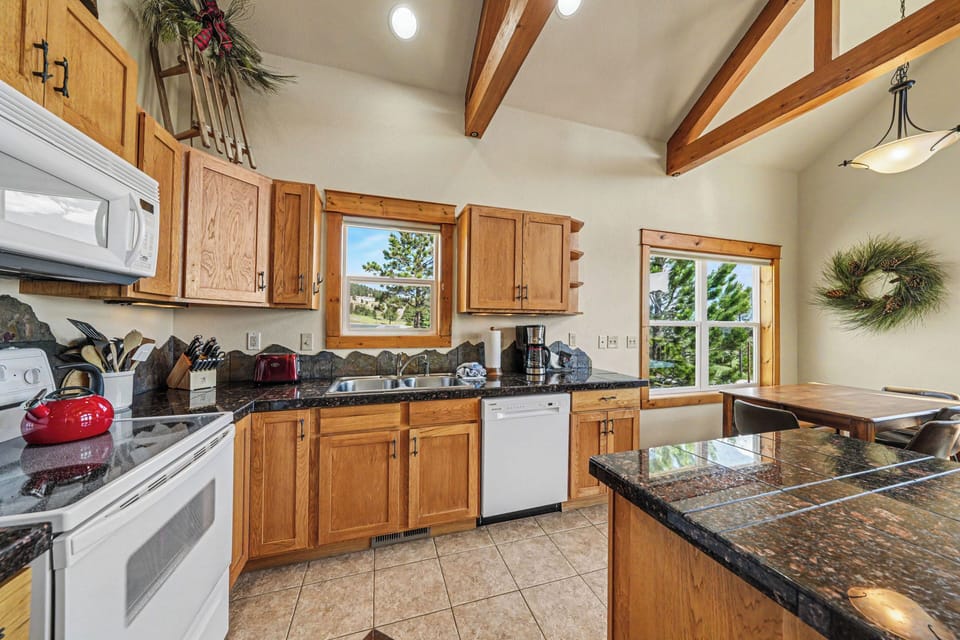 Your fully equipped kitchen features rich wood cabinetry, granite countertops, and vaulted ceilings with exposed beams, creating a warm and welcoming space to prepare meals with mountain views framing the windows.