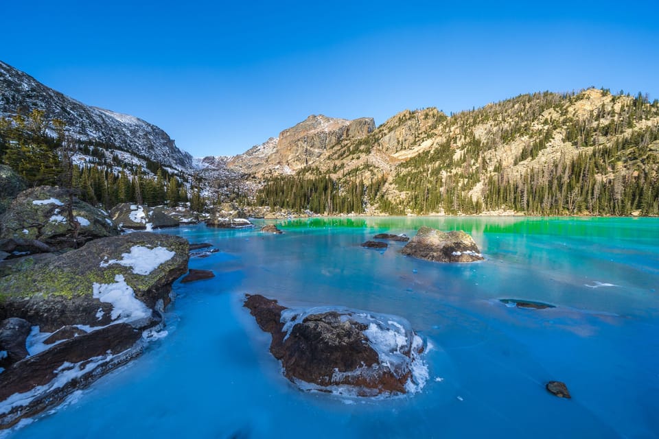 A serene mountain lake scene with turquoise water, surrounded by snowy rocks and forested mountains under a clear blue sky.