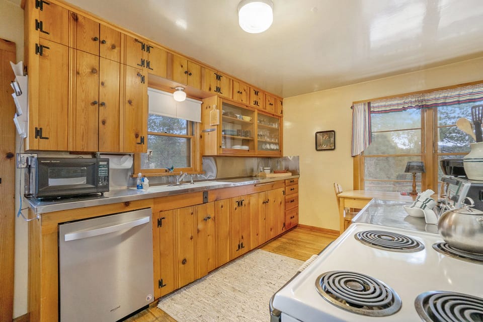 A wooden kitchen with retro-style cabinets, stainless steel appliances, a white electric stove, and large windows. The room is lit by ceiling lights.
