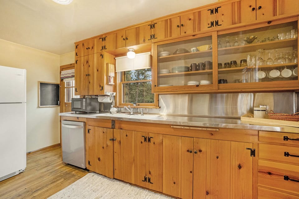 A kitchen with wooden cabinetry, a white refrigerator, a dishwasher, microwave, and a window above the sink. Upper cabinets have glass doors displaying dishes and glasses.