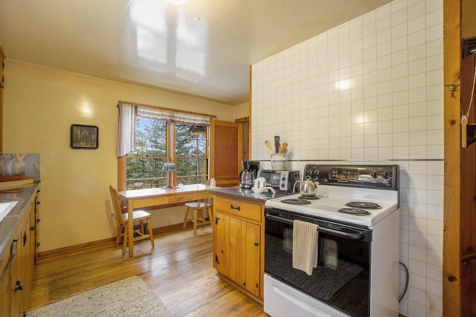 A cozy kitchen with wooden cabinets, an electric stove, and a window-side dining table with two chairs. Natural light streams in through the window.