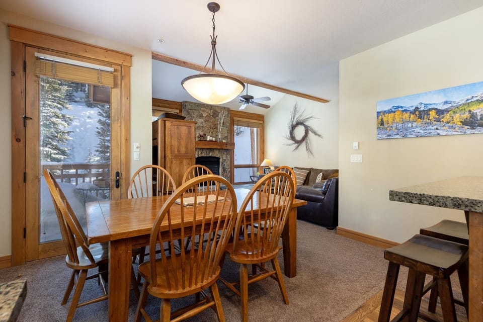 A dining area with a wooden table and six chairs, adjacent to a living room with a sofa, stone fireplace, and wall-mounted artwork. A door opens to a balcony with a snowy exterior view.