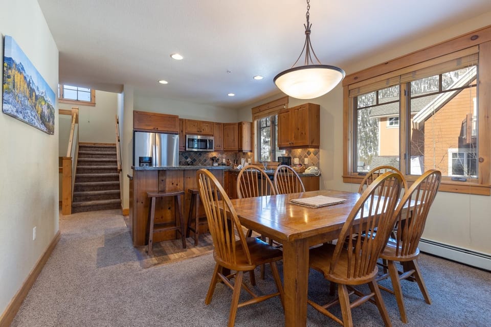 A dining area with a wooden table and eight chairs sits adjacent to a kitchen equipped with stainless steel appliances. A staircase is visible in the background, and a large window overlooks the exterior.