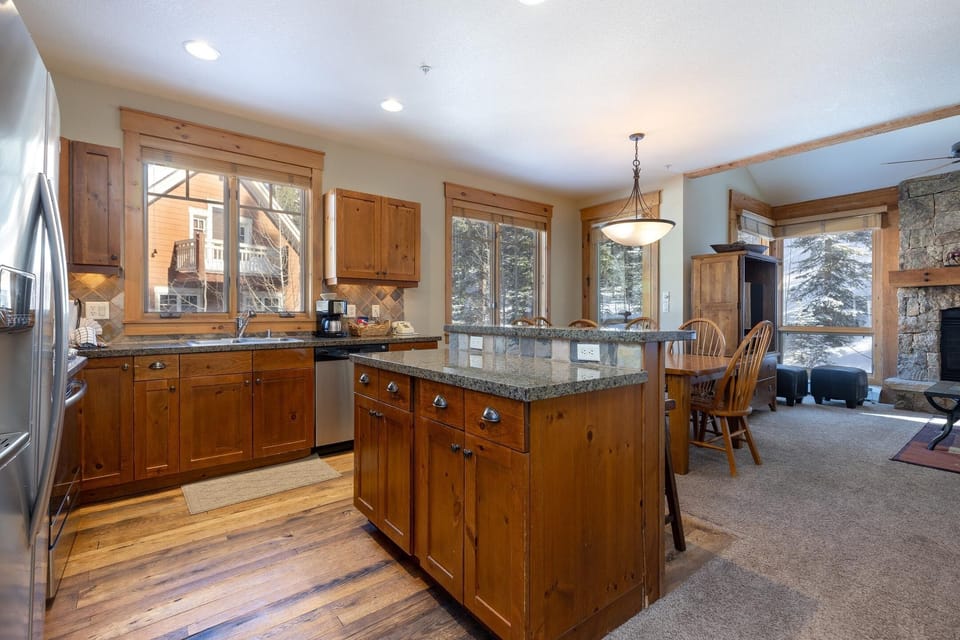 A rustic kitchen with wood cabinetry, granite countertops, an island, stainless steel appliances, and a dining area. Large windows offer natural light and a view of the outdoors.