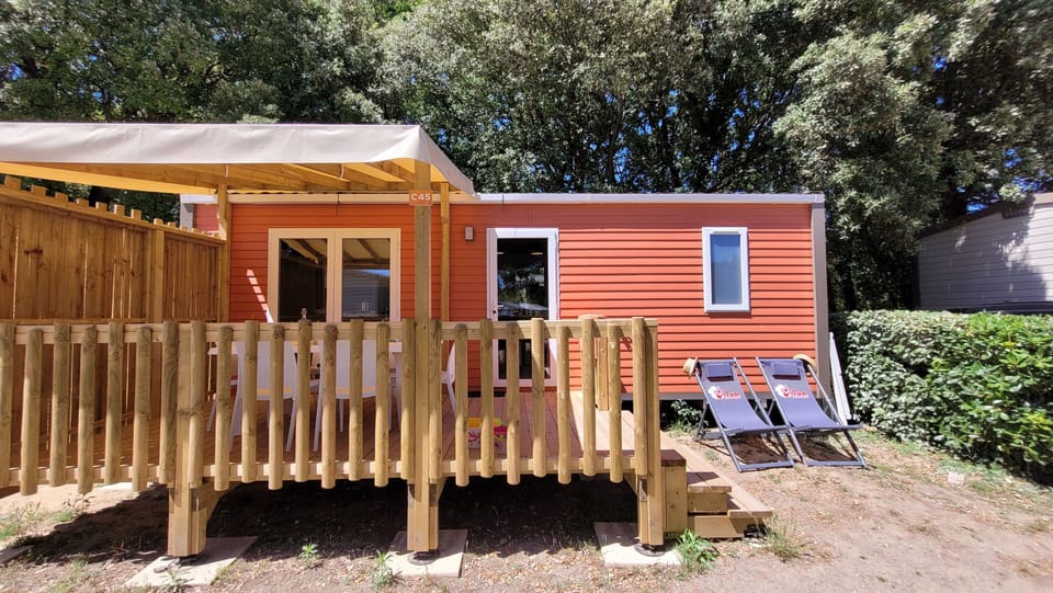 The vibrant exterior of the mobile home at Camping L’Orée du Bois showcases a cozy terrace with wooden railings, perfect for enjoying the outdoors.