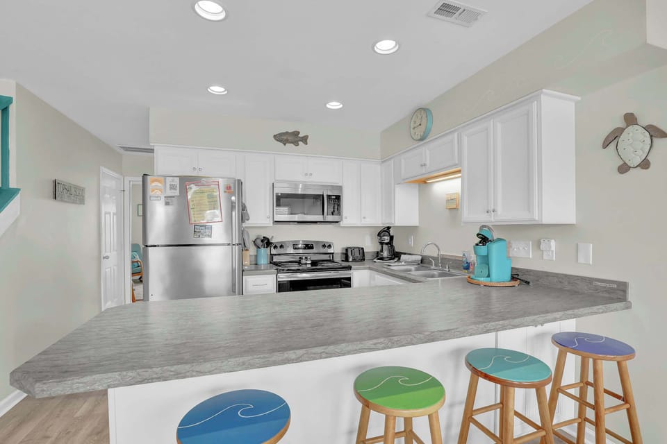 Everything you need for a week of beach cooking is right here — a fully equipped kitchen with stainless appliances, generous counter space, and wave-print bar stools that make morning coffee feel like it belongs on the shore.