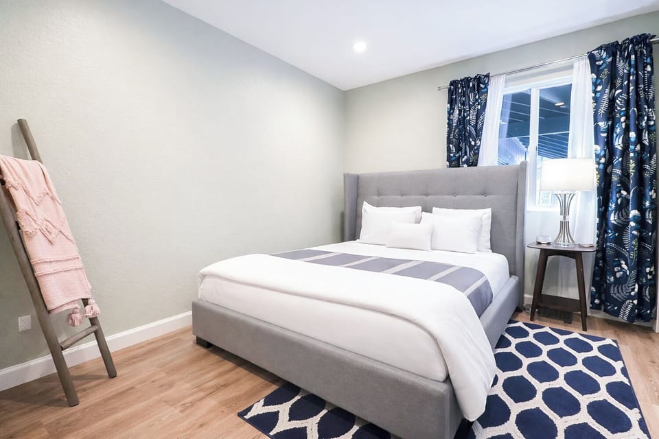 Sink into crisp white linens on your comfortable gray upholstered bed, surrounded by thoughtful touches and natural light streaming through tropical-print curtains.