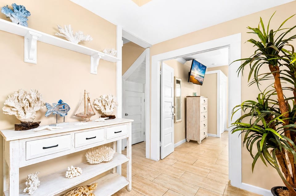 Coastal hallway with a white storage console, seashell and sailboat décor, indoor plant and doorway leading toward another bedroom.