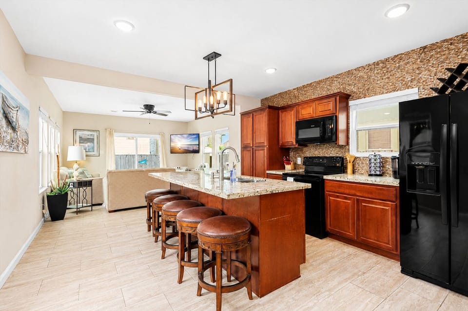 Open kitchen with granite breakfast bar, wood cabinetry, bar stools, black appliances and pendant lighting beside the coastal living area.