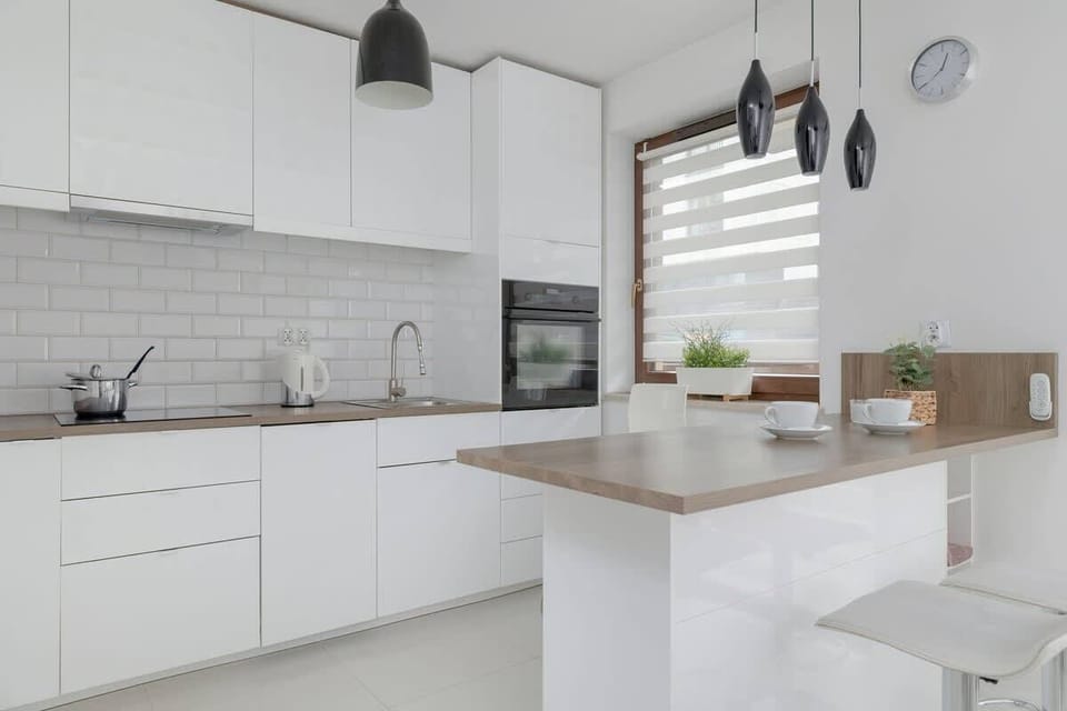 A sleek white kitchen with modern cabinets, a tiled backsplash, and a compact breakfast bar with two stools.

