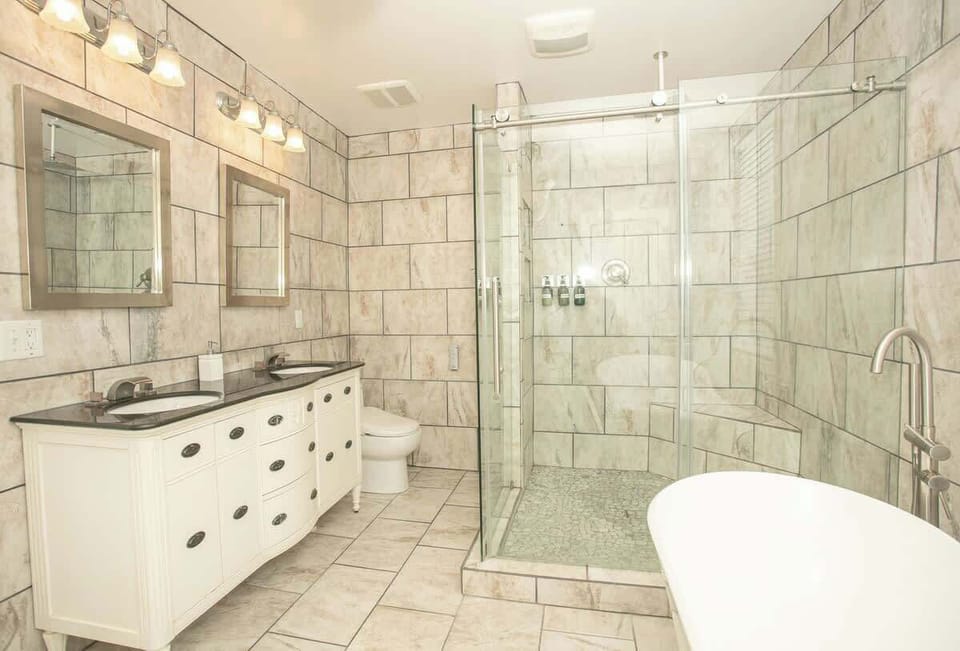 The spacious bathroom is tiled in marble and includes a dual vanity with black countertops, framed mirrors, a freestanding bathtub, and a large glass-enclosed shower with built-in shelving, all under bright vanity lighting.