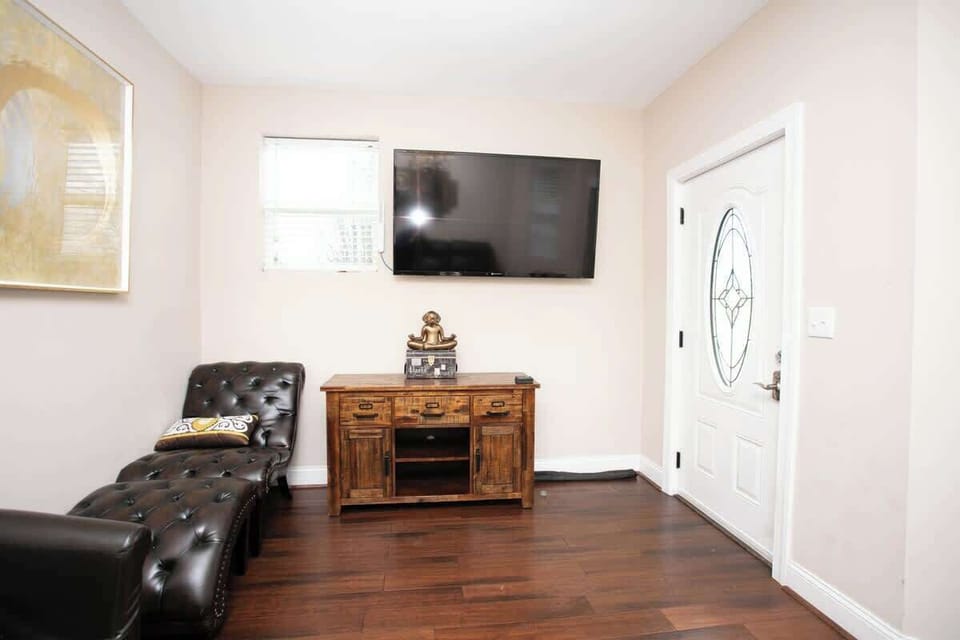 The living area features a tufted leather chaise lounge and a rustic console table beneath a wall-mounted flat-screen TV. A large abstract painting adds a touch of sophistication, while the entry door with decorative glass offers a warm welcome.