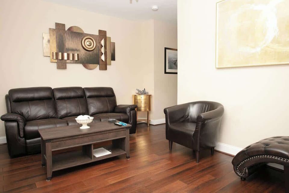 The living room is furnished with a comfortable dark leather couch and armchair, a wooden coffee table, and an ottoman. Abstract wall art and a gold accent table add elegance, while the rich wooden flooring provides warmth to the space.