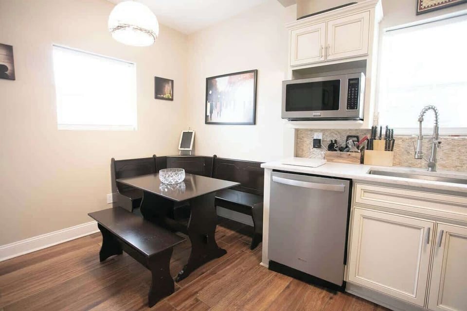 The kitchen dining space features a sleek, dark wood corner nook with a matching table under a chic pendant light. Cream cabinetry, a microwave, and modern appliances complement the mosaic backsplash and wooden flooring, creating a harmonious design.