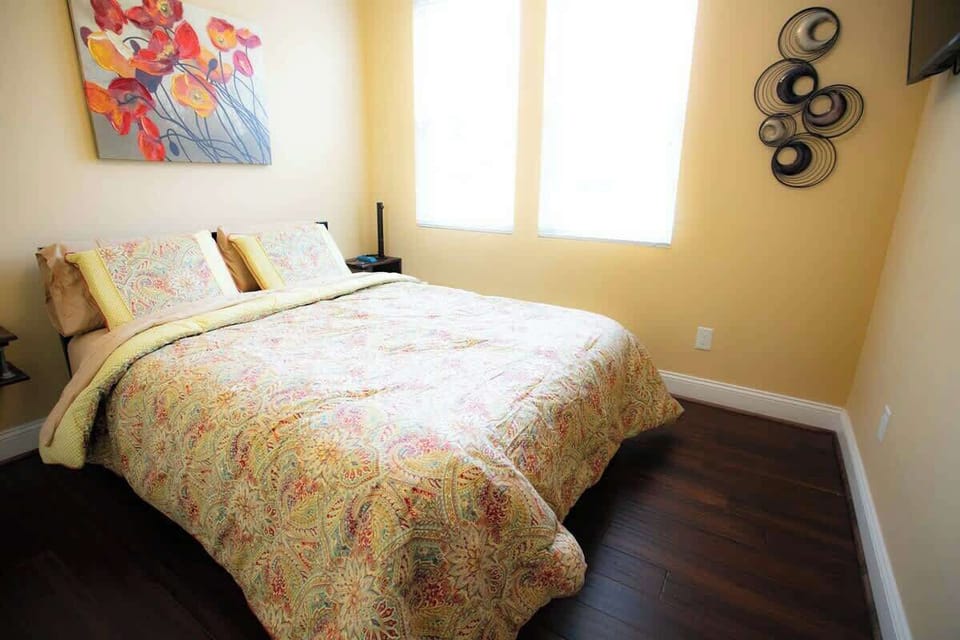 The bedroom is bright and cheerful, with a paisley-patterned comforter on the bed and a colorful floral painting above. Circular wall art adorns the adjacent wall, while natural light streams through two windows, highlighting the room's wooden floors