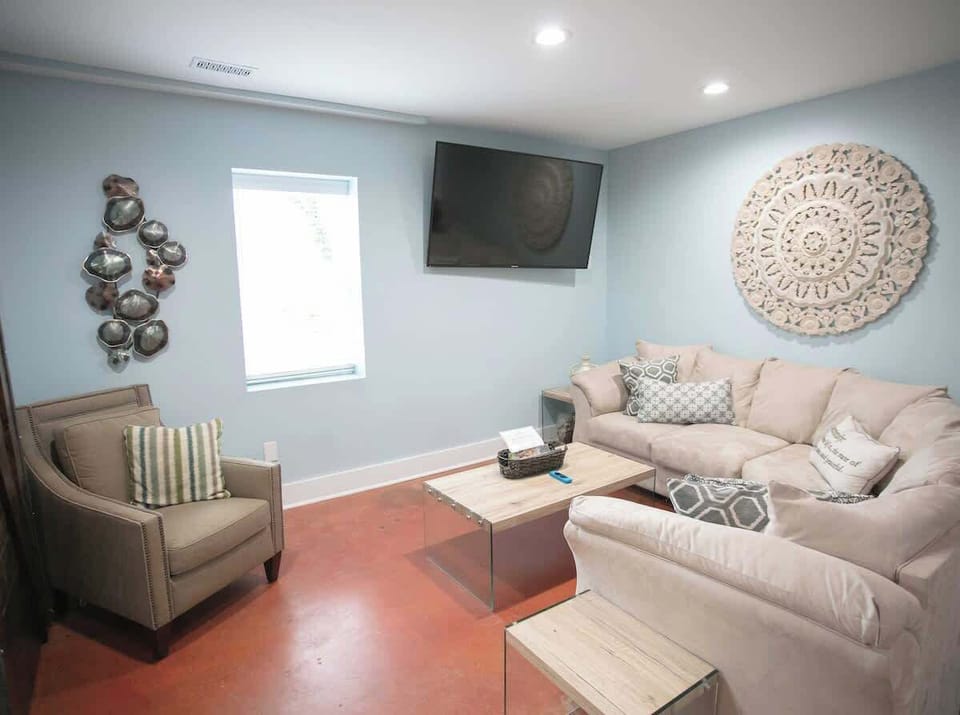 The living area is cozy with a large, comfortable sectional sofa adorned with decorative pillows. An intricate wall mandala, a rustic column, and a sleek wall-mounted TV contribute to the room's modern yet homely charm.