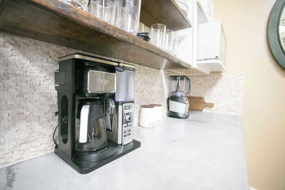 The kitchen counter is neatly organized with a modern coffee machine and a blender, ready for use. Above, open shelving displays a variety of glasses, showcasing a functional yet stylish setup.