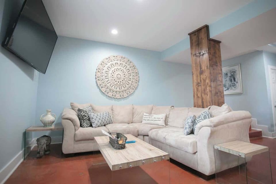 The living area is cozy with a large, comfortable sectional sofa adorned with decorative pillows. An intricate wall mandala, a rustic column, and a sleek wall-mounted TV contribute to the room's modern yet homely charm.