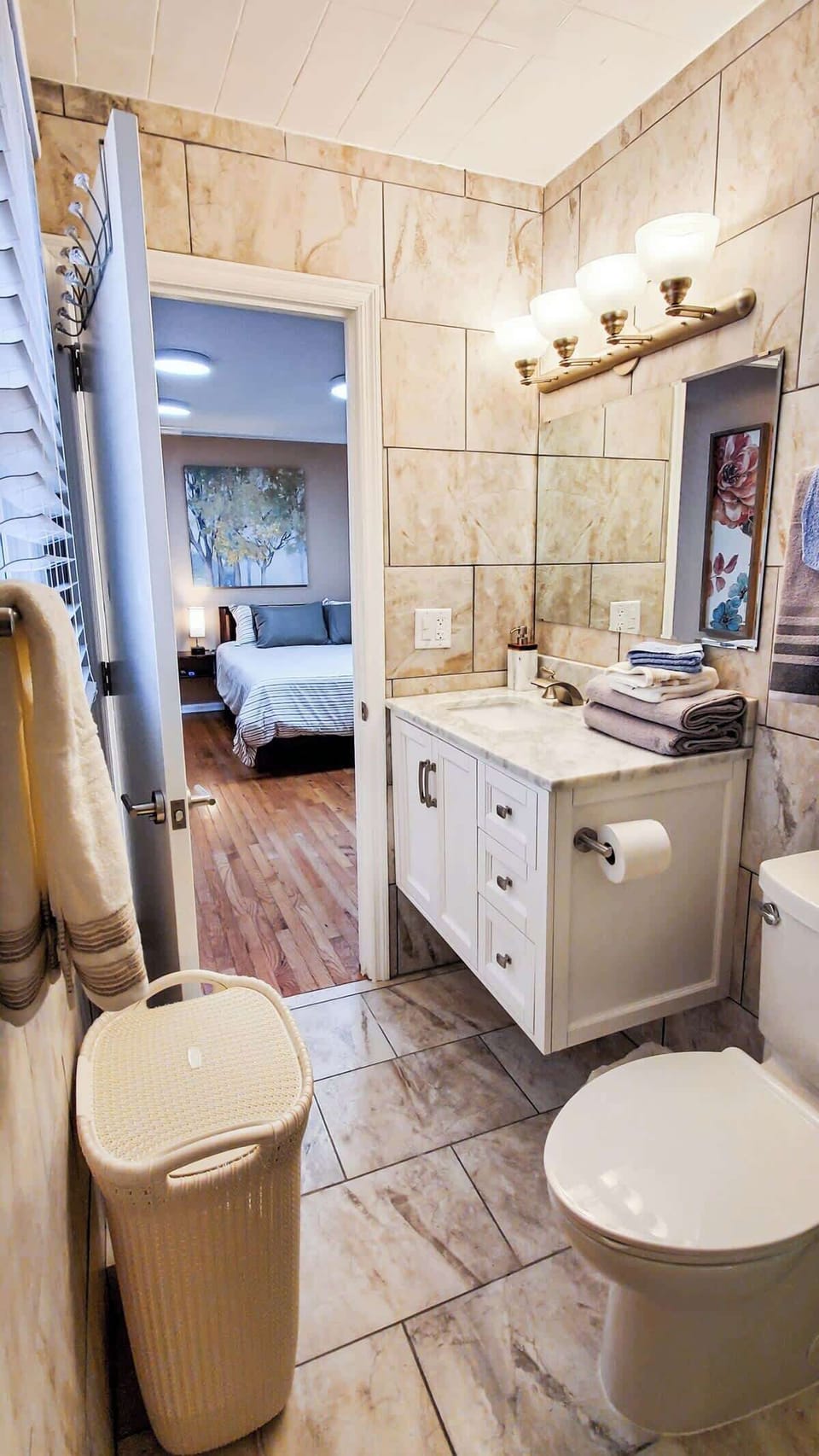 The bathroom is tastefully finished with marble tiles, a white vanity, and well-placed lighting, creating a sophisticated atmosphere. A view into the adjoining bedroom with hardwood floors reveals a cohesive, stylish interior design.