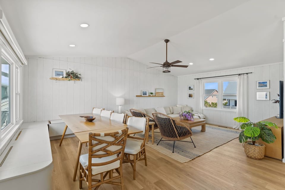 Dining room with a wood table, six chairs, white walls, coastal decor, and large windows framing the deck, white railings, and beautiful ocean views.