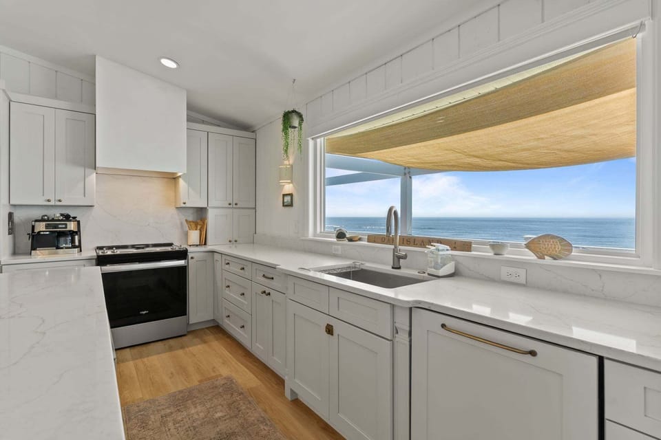Bright white kitchen with a large island, quartz counters, stainless appliances, farmhouse sink, and a big window overlooking the porch and ocean.