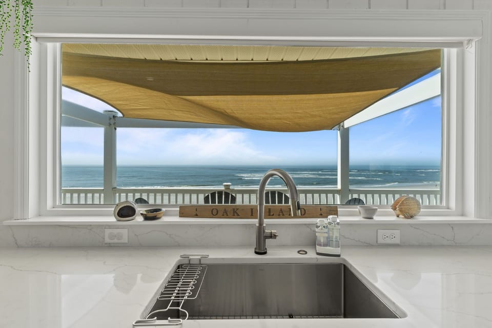 Bright white kitchen with a large island, quartz counters, stainless appliances, farmhouse sink, and a big window overlooking the porch and ocean.
