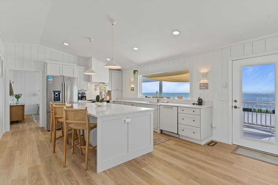 Bright white kitchen with a large island, quartz counters, stainless appliances, farmhouse sink, and a big window overlooking the porch and ocean.