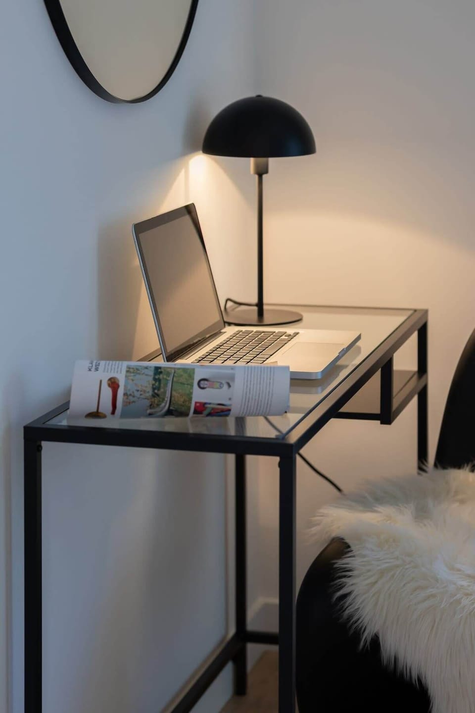 A bedside table with a black lamp, a book, and a fur-textured stool, creating a cozy reading corner.

