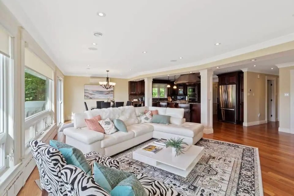 
A stylish living area with a plush white sofa, patterned chairs, and a chic rug, leading into a darker-toned dining space.
