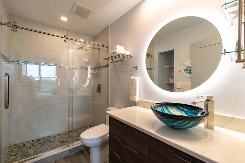 Modern bathroom featuring a glass shower and a distinctive blue vessel sink with backlit round mirror.