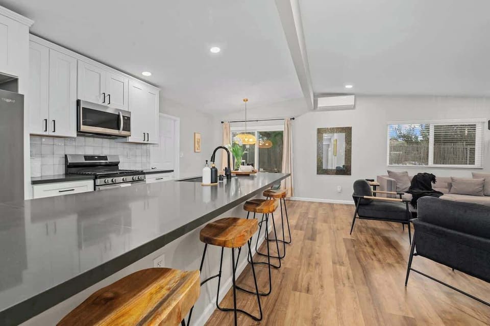 A bright, open-concept kitchen with white cabinetry, stainless steel appliances, and a sleek gray countertop with barstools. The space flows into a cozy living area with modern decor, wood flooring, and large windows.