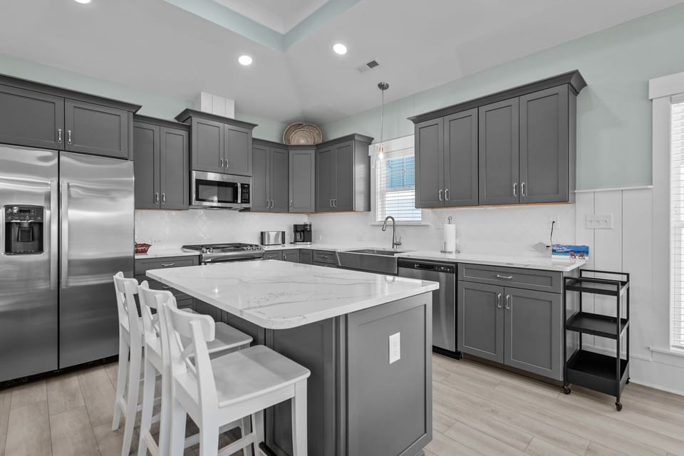 Bright kitchen with gray cabinets, stainless appliances, large island, white stools, farmhouse sink, dishwasher, microwave, coffee maker, toaster, and coastal Oak Island, NC style.