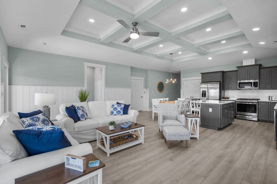 Bright coastal living room with white sofas, gray chairs, ottomans, wall TV, coffee table, blue pillows, tray ceiling, fan, tall windows, and beachy Oak Island, NC style.