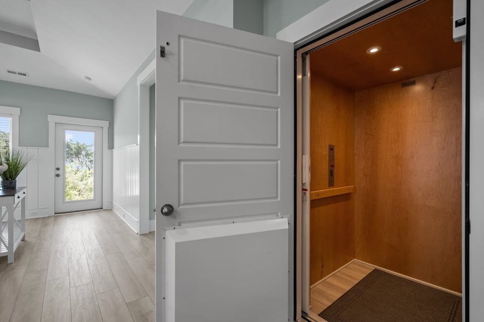 Convenient private elevator with bright coastal hallway, luxury vinyl plank flooring, white trim, and natural light. Accessible design adds comfort and ease to this spacious Oak Island, NC beach retreat.