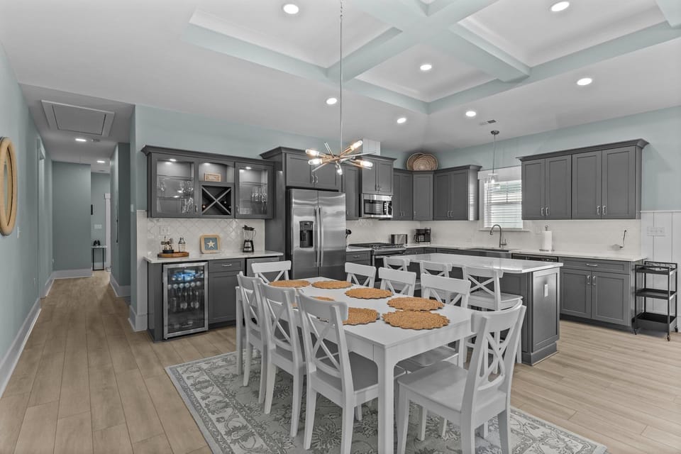Bright kitchen with gray cabinets, stainless appliances, large island, white stools, farmhouse sink, dishwasher, microwave, coffee maker, toaster, and coastal Oak Island, NC style.