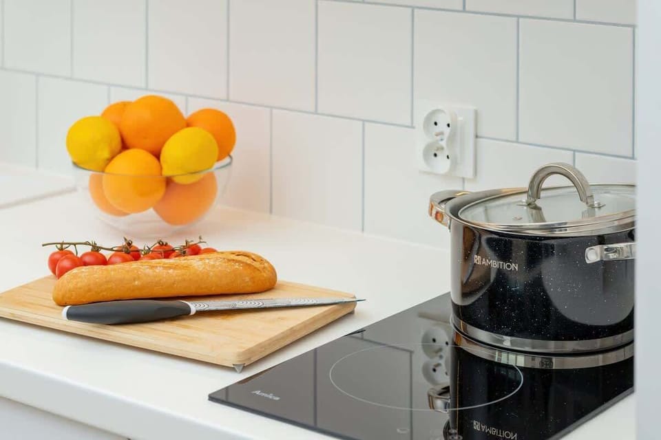 On the white kitchen worktop, against a wall of white tiles, lies a wooden board with a baguette and tomatoes, and next to it, on a black induction hob, stands a shiny black pot.